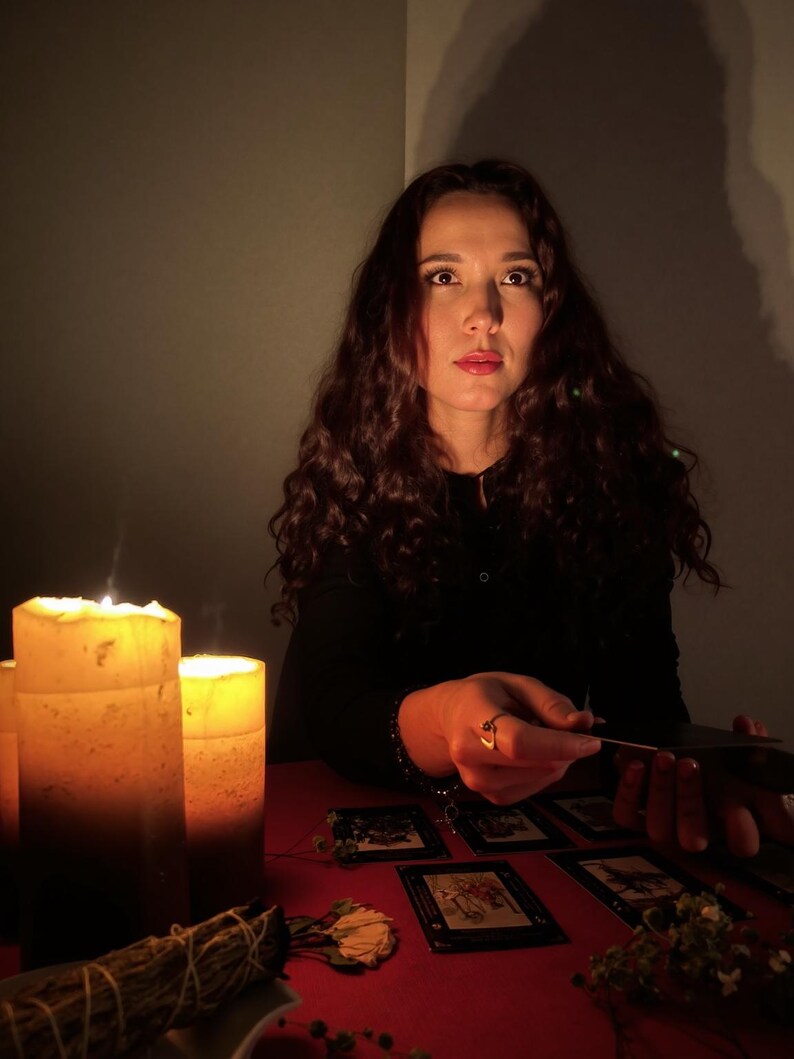 Puede incluir: Una mujer con largo cabello casta&ntilde;o rizado est&aacute; sentada en una mesa roja, iluminada por velas. Hay cartas del tarot dispuestas, y ella sostiene una. Un manojo de salvia y flores secas tambi&eacute;n est&aacute;n sobre la mesa, creando una atm&oacute;sfera m&iacute;stica.