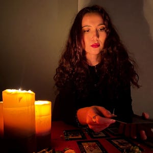 Pode incluir: Um conjunto de cartas de tarot dispostas sobre uma superf&iacute;cie vermelha com duas velas de pilar acesas que proporcionam uma ilumina&ccedil;&atilde;o quente. Um molho de s&aacute;lvia seca e pequenas flores secas est&atilde;o colocados perto das velas enquanto uma pessoa toca nas cartas.