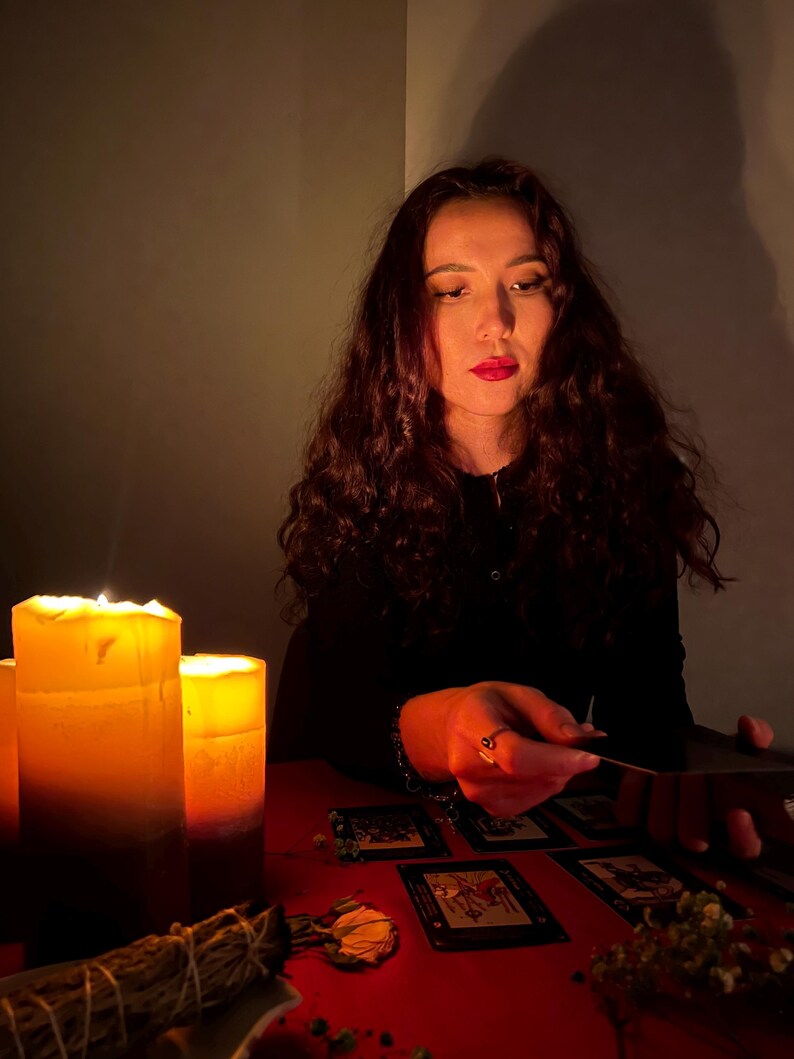 Pode incluir: Um conjunto de cartas de tarot dispostas sobre uma superf&iacute;cie vermelha com duas velas de pilar acesas que proporcionam uma ilumina&ccedil;&atilde;o quente. Um molho de s&aacute;lvia seca e pequenas flores secas est&atilde;o colocados perto das velas enquanto uma pessoa toca nas cartas.