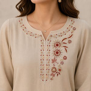 May include: A beige long-sleeved tunic featuring a split neckline with intricate embroidery. The design includes floral patterns in shades of deep red, gold, and white thread, concentrated along the neckline and extending down the center placket.