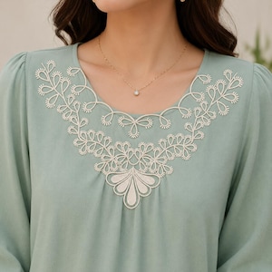 May include: A sage green long-sleeved blouse featuring a rounded neckline adorned with intricate cream-colored embroidery. The decorative stitching forms a symmetrical floral and vine pattern that extends across the chest area of the garment.