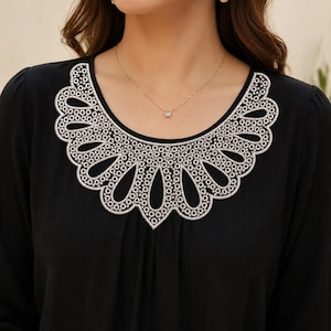 May include: A black long-sleeved shirt featuring a prominent white decorative collar with a scalloped edge and intricate cut-out petal patterns. The collar is detailed with a textured, beaded-style border that frames the neckline.