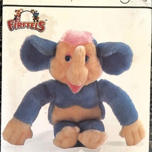 Op de afbeelding: Een Simplicity-knutselpatroon voor het maken van een pluche olifant en aap. Het patroon heet "Elephonkey" en toont een blauw en roze olifantenknuffel die op een witte achtergrond zit.