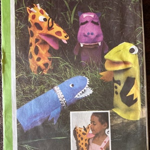 May include: A sewing pattern for making four finger puppets: a giraffe, a hippopotamus, a shark, and a frog. The pattern is numbered 8365. The puppets are shown in a green grassy setting. A child is shown using the giraffe puppet.