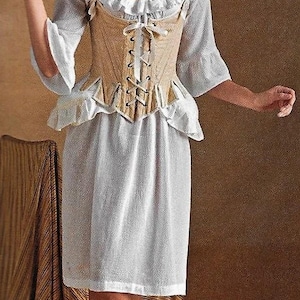 Könnte beinhalten: Ein weißes Kleid mit einem beigefarbenen Korsett-Oberteil und einem Rüschenkragen. Das Kleid hat ein tailliertes Oberteil und einen fließenden Rock. Das Model steht auf einer braunen und goldenen gestreiften Oberfläche.