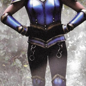 Puede incluir: Una mujer con armadura azul y dorada con un corpiño azul y pantalones a juego. La armadura tiene un detalle de cadena decorativo alrededor de la cintura. La mujer está de pie en un entorno de bosque.