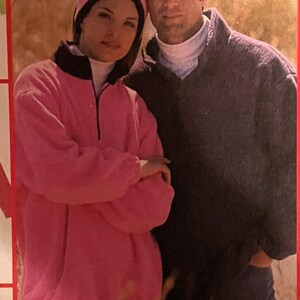 May include: A couple wearing casual clothing, standing in a field of tall grass. The woman is wearing a pink fleece jacket with a black zip and a black collar. The man is wearing a dark blue sweater. The text "GREAT LOOKS 1 EASY PATTERN" is visible at the top of the image.