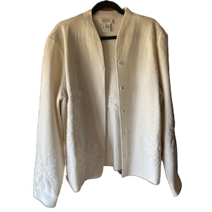 May include: A cream-coloured long-sleeve cardigan featuring a V-neckline and a button-front closure. The garment is decorated with tonal floral embroidery on the lower front panels and sleeves, and includes a brand label at the inner neckline.