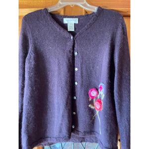 May include: A dark purple long-sleeve cardigan with a V-neckline and a button-down front. The garment features a decorative embroidered floral design with pink and red fabric roses on the lower right side and a scalloped edge along the neckline.
