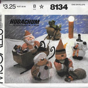 May include: A vintage McCall's sewing pattern envelope featuring a "Hugachum" design. The image showcases handmade Christmas-themed plush figures, including Santa Claus in a sleigh, a reindeer, a snowman, and other characters.