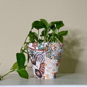 May include: A decorative flower pot with a patchwork design of paisley and floral patterns in shades of white, blue, orange, and brown. The pot contains a green leafy plant with trailing vines.