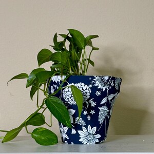 May include: A potted plant with green leaves in a navy blue flower pot. The pot is decorated with a white floral pattern. The plant is on a white shelf against a beige wall.