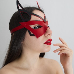 Op de afbeelding: Een rood en zwart leren masker met een kattenoogontwerp. Het masker heeft een rode oogbedekking met zwarte accenten en een bijpassende hoofdband. Het masker wordt op het gezicht gedragen.