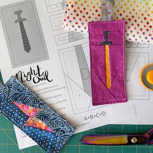 May include: A close-up of a pink fabric pouch with a yellow and black sword design. The pouch is lying on a white sheet of paper with instructions for a sewing project. The paper includes the text "Night Owl Quilts" and "A+B+C+D".