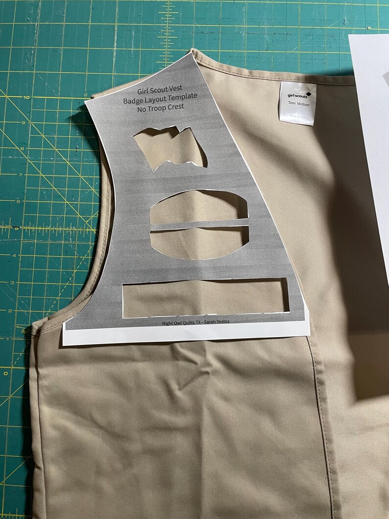 May include: A tan Girl Scout vest with a printed badge layout template. The template includes spaces for badges, a troop crest, and a Girl Scout logo. The text on the template reads "Girl Scout Vest Badge Layout Template No Troop Crest" and "Night Owl Quilts TX - Sarah Yates".