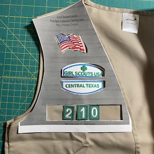 May include: A tan Girl Scout vest with a badge layout template. The template includes a space for the American flag, a Girl Scouts USA patch, a Central Texas patch, and a number patch. The text "Girl Scout Vest Badge Layout Template No Troop Crest" is printed on the template. The text "girl scouts Tem Medium" is printed on the vest.