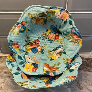 May include: Two quilted bowl cozies with a teal background and a colorful bird and floral pattern. The cozies are quilted with a diamond pattern. The birds are red, blue, yellow, and white. The flowers are pink, yellow, and orange.