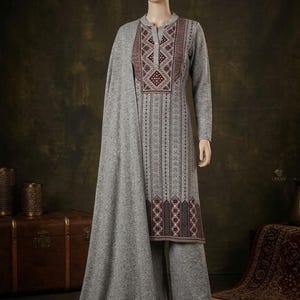 Op de afbeelding: Een grijze tuniek met lange mouwen, een opstaande kraag en een knoopsluiting, voorzien van verticale strepen en geometrisch borduurwerk in bordeauxrood en wit. De tuniek wordt gecombineerd met een bijpassende wijde broek en een lange, effen grijze sjaal.