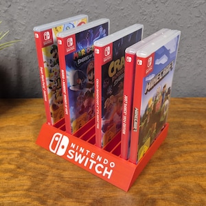 Puede incluir: Un soporte rojo para juegos de Nintendo Switch con cuatro estuches de juegos. El soporte tiene el logotipo de Nintendo Switch en blanco. Los estuches de juegos tienen gráficos coloridos y títulos como "Minecraft" y "Mario + Rabbids Sparks of Hope".