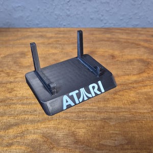 May include: Black plastic Atari game console stand with the Atari logo in light blue. The stand has two vertical supports and sits on a wooden surface.