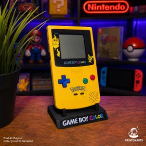 Op de afbeelding: Een gele Game Boy Color handheld spelcomputer met Pikachu-afbeeldingen op de schermrand, een blauwe richtingsknop, groene en rode knoppen en het Pokémon-logo, geplaatst op een zwarte standaard met de tekst Game Boy Color erop.