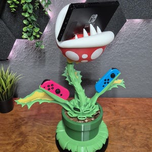 Op de afbeelding: Een 3D-geprinte Nintendo Switch-standaard in de vorm van een Piranha Plant uit de Super Mario-serie. De standaard is rood, wit en groen, met sleuven voor de Switch-console en Joy-Con controllers. Hij staat op een houten oppervlak.