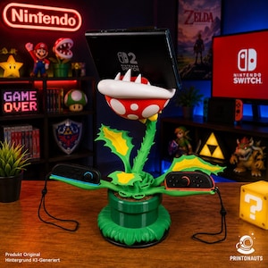 Könnte beinhalten: Ein grüner Piranha-Pflanzen-Gaming-Zubehörständer mit einem rot-weiß gepunkteten Kopf, der eine Nintendo Switch-Konsole hält, sowie zwei grünen blattförmigen Armen für Joy-Con-Controller, alles auf einer grünen Röhrenbasis montiert.