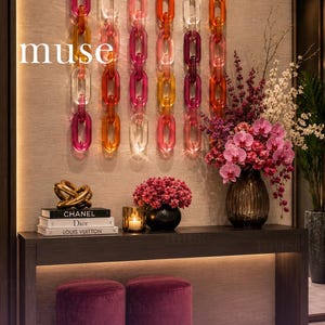 Peut inclure: Une décoration murale composée de six chaînes verticales en acrylique translucide aux tons rose, orange et transparent. En dessous, une console sombre supporte trois livres de mode empilés, une sculpture dorée, une bougie et un vase de fleurs.