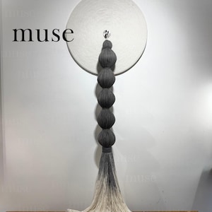 Könnte beinhalten: Ein Wandbehang mit einer großen weißen kreisförmigen Scheibe mit konzentrischer Textur. Eine vertikale Säule aus fünf dunkelgrauen Garnkugeln hängt in der Mitte und geht in eine lange, fließende Ombré-Quaste über, die von Dunkelgrau zu Weiß verläuft.