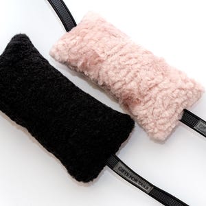 Könnte beinhalten: Zwei rechteckige, flauschige und strukturierte Abdeckungen für Riemen, eine in Schwarz und eine in Hellrosa. Jede Abdeckung ist an einem schwarzen elastischen Riemen befestigt, der ein kleines graues rechteckiges Etikett mit dem Text GIFTS FOR TAILS trägt.