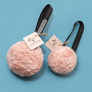 May include: Two pink dog toys with black handles and tags reading "GIFTS FOR TAILS" against a light blue backdrop. The toys are round and fluffy, with a soft texture, ideal for pets.