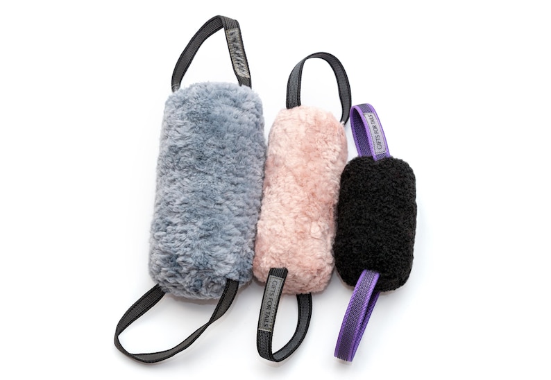 Pu&ograve; includere: Tre giocattoli di peluche per cani in grigio, rosa e nero. Ogni giocattolo ha un'impugnatura ad anello e un'etichetta con la scritta "GIFTS FOR TAILS". I giocattoli sono cilindrici e realizzati in un materiale morbido e strutturato.