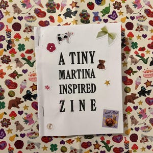 May include: A white zine titled "A Tiny Martina Inspired Zine" with decorative stickers of a cow, bear, flower, bow, and a postage stamp. The zine is on a patterned background with various stickers.