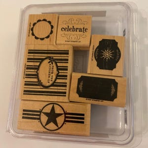 May include: A collection of wooden stamps with various designs and text, including "celebrate", "From the Kitchen of", and "winter wishes". The stamps are in a clear plastic container. The stamps have black ink designs.