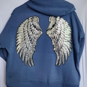 May include: A blue hooded sweatshirt with silver sequined angel wings on the back. The wings are detailed with individual sequin feathers. The sweatshirt has a drawstring hood and a ribbed hem.