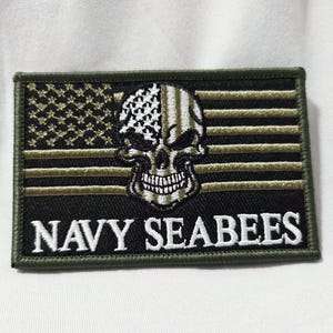 May include: A rectangular embroidered patch featuring a stylized skull centered over a black and olive green American flag design. The text NAVY SEABEES is stitched in white capital letters across the bottom portion of the patch border.