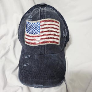 May include: A distressed navy blue baseball cap featuring a sequined American flag patch. The flag design includes red and white stripes and a blue field with white stars. The cap has a curved brim and a button on top.