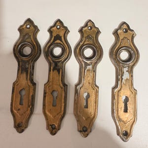 Puede incluir: Cuatro embellecedores de puerta antiguos de latón con cerraduras y detalles decorativos. Los embellecedores tienen un acabado dorado deslustrado con signos de desgaste y edad. Los agujeros de la cerradura son ovalados.