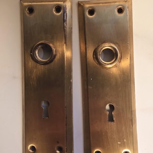 Könnte beinhalten: Zwei antike Türschilder aus Messing. Jede Platte hat ein Schlüsselloch, eine kreisförmige Öffnung und vier Schraubenlöcher. Das Messing hat eine warme, gealterte Patina mit Abnutzung und Verfärbung. Die Platten sind rechteckig.