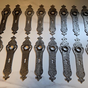 Peut inclure: Une collection de poignées de porte anciennes en métal. Chaque poignée présente un design orné et allongé avec un trou de serrure et une ouverture de poignée circulaire. Le métal a un aspect texturé et vieilli, suggérant une esthétique vintage. Les poignées sont disposées sur une surface claire.