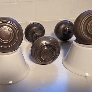 May include: Five antique bronze door knobs with a circular design, displayed on white ceramic stands. The knobs have a dark, aged appearance, suggesting they are vintage or reclaimed. The arrangement highlights the knobs' intricate details.