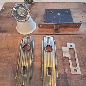Peut inclure: Un ensemble de quincaillerie de porte ancienne comprenant un bouton en verre transparent avec une base en porcelaine blanche, une serrure à mortaise en métal noir, deux plaques en laiton rectangulaires à rainures verticales et une gâche.