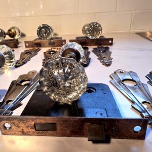 Pode incluir: Coleção de puxadores de porta de vidro vintage e ferragens. Os puxadores são transparentes, facetados e redondos, com componentes de latão e metal. As ferragens incluem placas e fechos de metal, mostrando uma estética antiga.