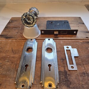 May include: A set of vintage door hardware including a clear glass knob with a white base, a black metal mortise lock box, two rectangular silver-toned metal backplates with keyholes, and a small silver-toned metal strike plate for a door.