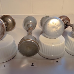 May include: Four antique door knobs in various styles. The knobs are made of metal and have different finishes, including tarnished silver and dark brown. Each knob is placed on a white ceramic base with a number label.