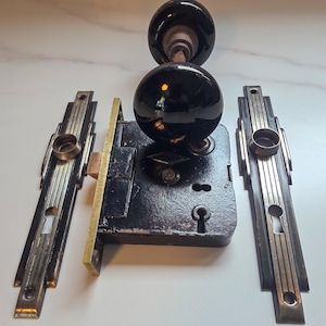 Peut inclure: Un ensemble de serrure de porte ancien avec un bouton de porte noir et rond et une plaque arrière assortie. L'ensemble comprend un mécanisme de verrouillage noir et deux rosaces rectangulaires décoratives avec des trous de serrure.