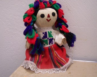 May include: A handmade rag doll with colorful yarn hair, a white dress with embroidered details, and a red skirt with colorful stripes. The doll is wearing a white lace trim.
