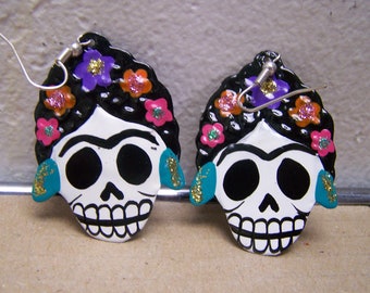 May include: A pair of silver dangle earrings featuring a white skull with black eyes and a black painted hairstyle. The skull is decorated with colorful flowers and glitter.
