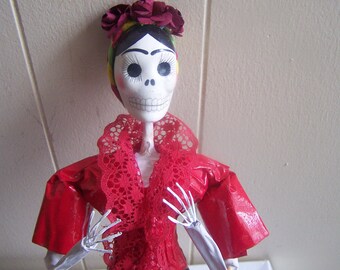 May include: A white skeletal doll with a red and white lace collar, wearing a red dress and a red flower crown.