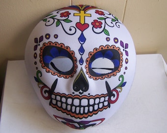 May include: A white plastic Day of the Dead mask with colorful floral and skull designs. The mask has blue eyes, a black nose, and a red heart on the forehead.